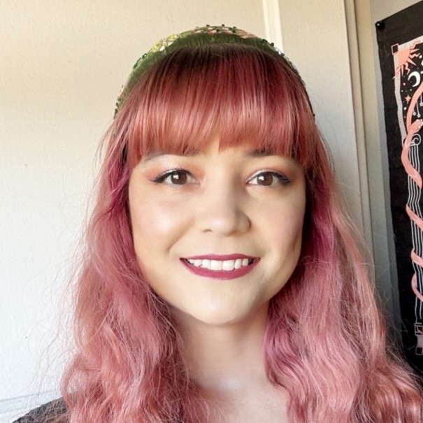 a photo of Cassandra Garcia. A smiling woman with dyed dusky pink hair, and lipstick.