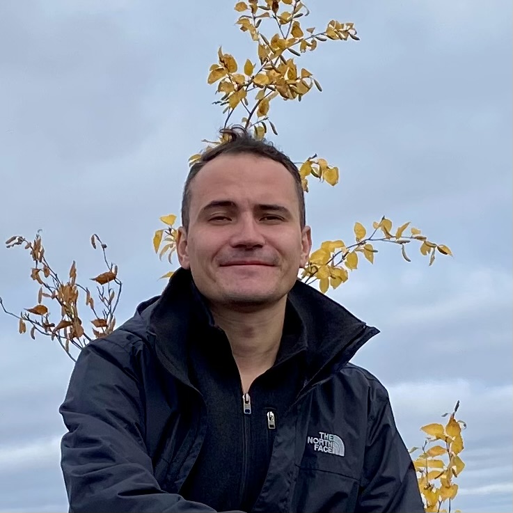 Image of a man in a black jacket smiling, standing outdoors in front of yellow autumn leaves and a cloudy sk