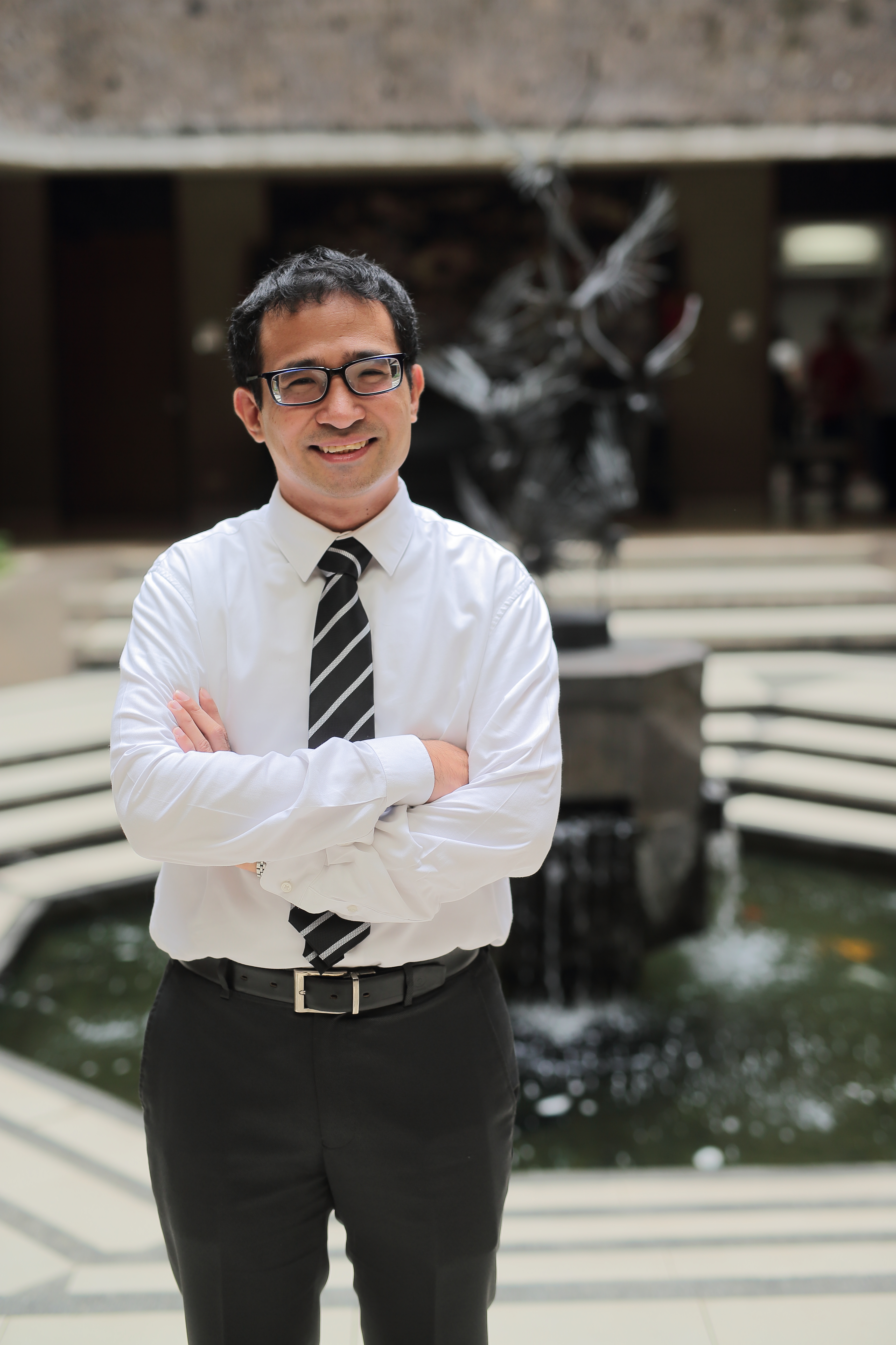 Varsolo Sunio A man in glasses and a white shirt with a striped tie stands confidently with arms crossed in front of a circular water fountain