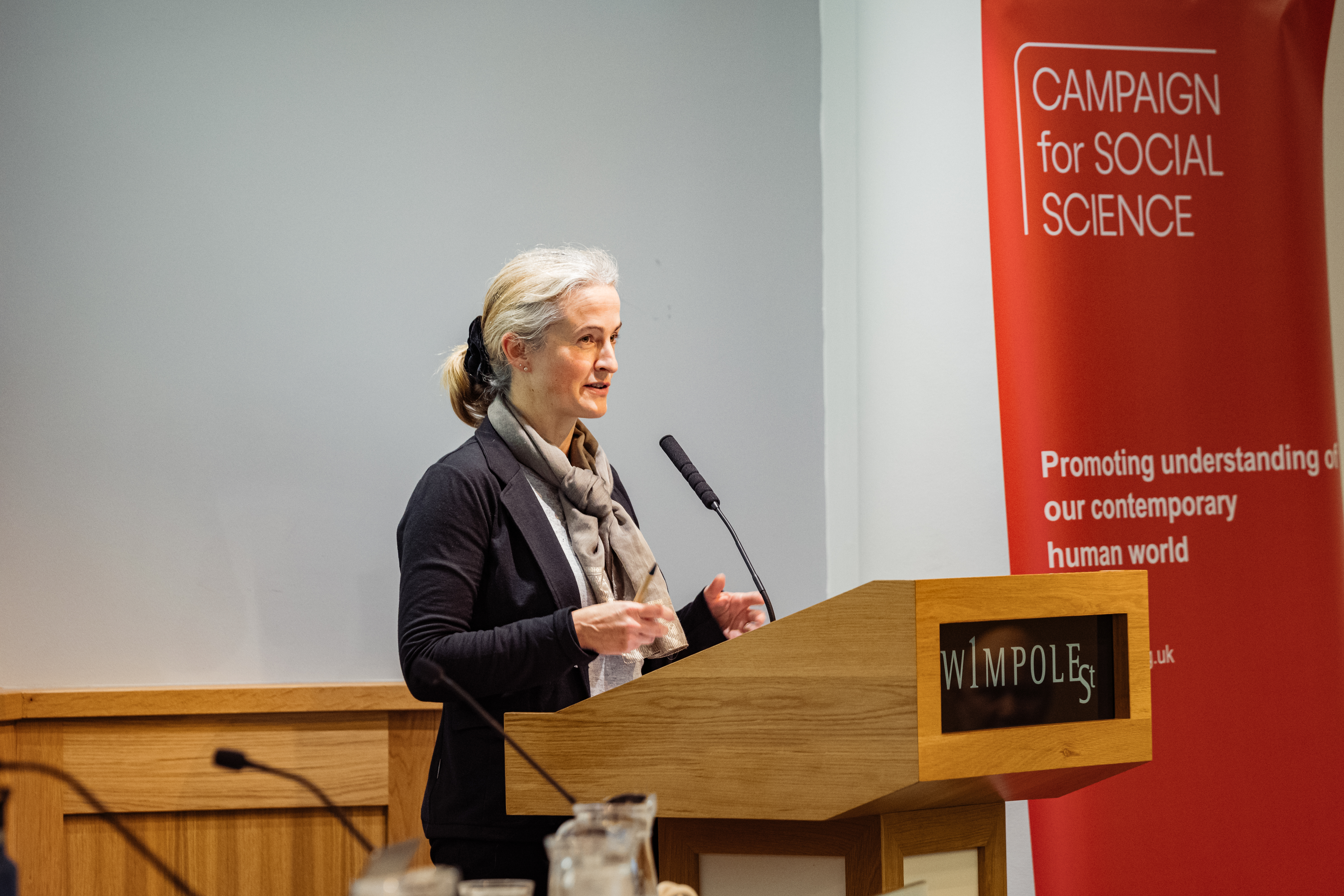 Dr. Hannah White standing at a lectern speaking into the attached microphone. Campaign for Social Science banner is to the right.