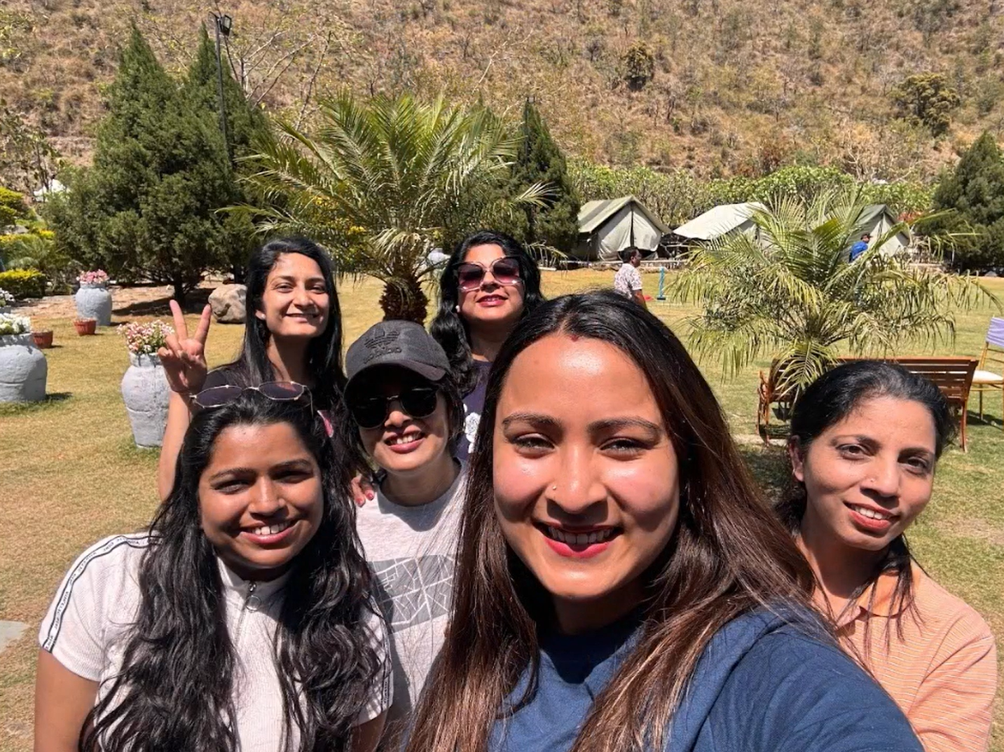 Six women smiling with plants, trees, and flower pots  behind them.