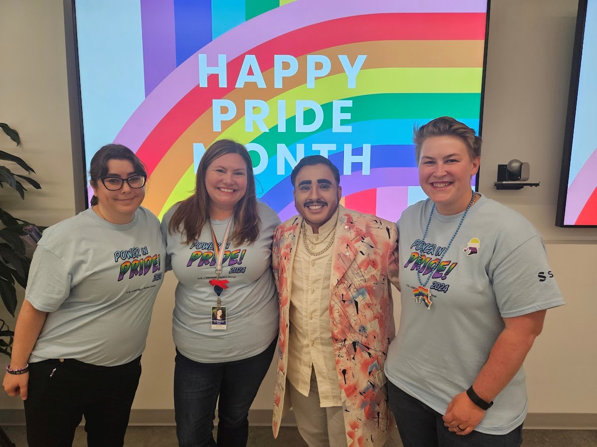 Four people standing next to each other and smiling, with "HAPPY PRIDE MONTH" and rainbow colors on a screen behind them