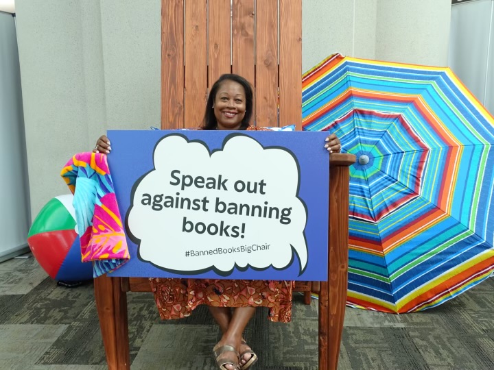 Sonja Cherry-Paul sitting on an oversized chair  with a beach umbrella to the right and a beach ball to the left. She's holding uo a sign that reads "Speak out against bannking books! #BannedBooksBigChair"